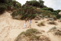 Восхитительный многорасовый мост и жених, держащиеся за руки и бегущие вдоль песчаного холма в день свадьбы на природе — стоковое фото