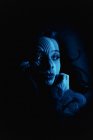 Mujer joven y soñadora con luz y sombra a rayas en la cara mirando hacia otro lado pensativamente mientras yace en la oscuridad - foto de stock