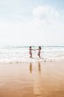 Vista lateral de amigas alegres de mãos dadas em fatos de banho no oceano espumoso perto da praia de areia sob o céu azul nublado no dia ensolarado — Fotografia de Stock