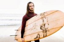 Homem surfista vestido de fato de mergulho a caminhar com a prancha de surf em direcção à água para apanhar uma onda na praia durante o nascer do sol — Fotografia de Stock