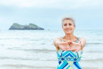 Active old gray haired female pensioner in stylish swimsuit stretching arms and shoulders while doing exercises on beach against waving ocean — Stock Photo