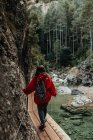 Visão traseira do jovem turista com mochila andando na passarela acima da água perto da parede de pedra entre a floresta — Fotografia de Stock