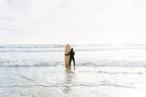 Обратный вид на неузнаваемого серфера, одетого в гидрокостюм, стоящего, смотрящего с доской для серфинга в сторону воды, чтобы поймать волну на пляже во время восхода солнца — стоковое фото