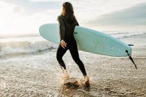 Vue latérale de surfeur homme vêtu d'une combinaison de plongée marchant avec planche de surf vers l'eau pour attraper une vague sur la plage au lever du soleil — Photo de stock