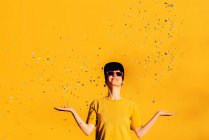 Feminino moderno em hip hop cap e óculos de sol soprando confete colorido e se divertindo contra o fundo amarelo — Fotografia de Stock