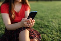 Weibchen in Sommerkleidung sitzen auf der grünen Wiese im Park und surfen mit dem Handy im Internet und unterhalten sich am Wochenende abends — Stockfoto