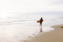 Vista trasera de un surfista irreconocible vestido con traje de neopreno que huye con la tabla de surf hacia el agua para atrapar una ola en la playa durante el amanecer - foto de stock