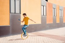 Cuerpo completo de hombre joven ágil que utiliza el teléfono móvil mientras monta monociclo cerca de un edificio urbano colorido moderno - foto de stock