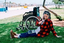 Cuerpo completo de mujer de pelo gris positivo con computadora portátil en las rodillas respondiendo a la llamada telefónica mientras está sentado en la hierba verde cerca de la silla de ruedas en el parque - foto de stock