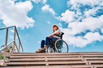 Низький кут повного тіла жінки з інвалідністю сидить у інвалідному візку біля сходів, дивлячись на блакитне небо в місті — стокове фото