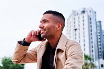 Low angle of positive well dressed young Hispanic businessman talking on smartphone and discussing news on urban street with contemporary buildings in background — Stock Photo