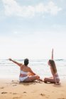 Back view of unrecognizable smiling young female friends in white swimsuits sitting on sandy shore near ocean under blue cloudy sky in sunny day — Stock Photo