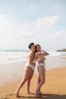 Side view of cheerful female friends in swimsuits embracing each other while standing in foamy ocean near sandy beach under blue cloudy sky in sunny day — Stock Photo