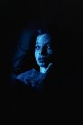 Mujer joven y soñadora con luz y sombra a rayas en la cara mirando hacia otro lado pensativamente mientras yace en la oscuridad - foto de stock