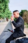 Vista lateral de bajo ángulo del joven hispano en traje negro formal con teléfono móvil en la mano disfrutando de refrescante bebida para llevar mientras descansa en la calle urbana en el día de verano - foto de stock