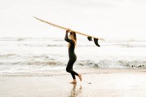 Vue latérale d'une surfeuse vêtue d'une combinaison de plongée regardant loin tout en tenant une planche de surf sur la tête sur la plage au lever du soleil en arrière-plan — Photo de stock