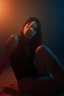 Junges, emotionsloses weibliches Modell mit kreuzförmiger Lichtprojektion auf das Gesicht, das im dunklen Studio sitzt und in die Kamera blickt — Stockfoto