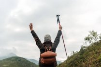 Back view of anonymous aged woman with backpack holding walking stick in raised arms against cloudy gray sky while exploring nature — Stock Photo