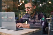 Sério gerente masculino hispânico em camisa violeta e óculos digitando no teclado netbook enquanto sentado à mesa atrás da janela e trabalhando no projeto no café — Fotografia de Stock