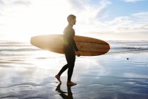Vue latérale de surfeur homme vêtu d'une combinaison de plongée marchant sur la plage avec la planche de surf le matin avec lever de soleil en arrière-plan — Photo de stock
