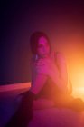 Tranquille jeune mannequin femme en robe assise sur le sol et penchée sur le genou tout en regardant loin dans un studio sombre avec des lumières colorées — Photo de stock