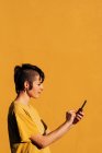 Vue latérale de la femme contemporaine avec coupe de cheveux élégante et perçage à l'aide d'un smartphone pour envoyer un message texte dans les médias sociaux sur fond jaune — Photo de stock