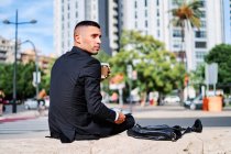Низкий угол зрения серьезный задумчивый молодой латиноамериканец в формальном черном костюме задумчиво глядя в сторону, сидя с цифровым гаджетом в руке на городской улице — стоковое фото
