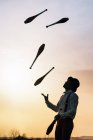 Низкий угол обзора неузнаваемого циркового артиста в шляпе жонглирует клубами на закатном небе — стоковое фото