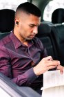Hombre de negocios hispano serio con verdaderos auriculares inalámbricos leyendo notas en un cuaderno mientras está sentado en el asiento trasero del automóvil y viajando al trabajo - foto de stock