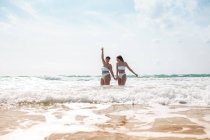 Снизу веселые подруги держатся за руки в купальниках в пенном океане возле песчаного пляжа под голубым облачным небом в солнечный день — стоковое фото
