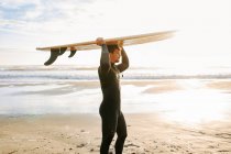 Vista laterale dell'uomo surfista vestito di muta che cammina sulla spiaggia con la tavola da surf sopra la testa al mattino con l'alba sullo sfondo — Foto stock