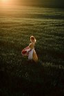 Молодая женщина в винтажном стиле платье с плетеной корзиной во время прогулки по зеленому травянистому полю на закате в летнее время в сельской местности — стоковое фото