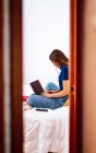 Vista lateral de una joven freelancer positiva en ropa casual sentada con las piernas cruzadas en la cama y escribiendo en el portátil mientras trabaja en un proyecto remoto en casa - foto de stock
