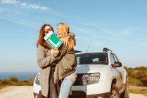 Cheerful young girlfriends with Low gear sign standing at white car on shore of sea and embracing having trip — Stock Photo