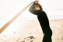 Seitenansicht eines lächelnden Surfers im Neoprenanzug, der mit dem Surfbrett am Strand bei Sonnenaufgang im Hintergrund in die Kamera schaut — Stockfoto