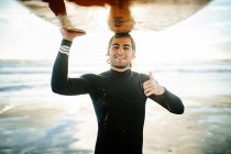 Retrato del joven surfista feliz vestido con traje de neopreno de pie con los pulgares hacia arriba mirando la cámara en la playa con la tabla de surf por encima de la cabeza durante el amanecer - foto de stock