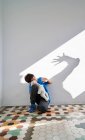 Scared little boy with toy in hands sitting near wall with shadow of angry violent parent with raised arm — Stock Photo