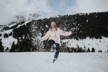 Joven turista feliz en gafas con la pierna levantada mirando a la cámara y divirtiéndose entre el campo en la nieve cerca de la montaña - foto de stock