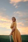 Vista lateral de la mujer en vestido de estilo vintage de pie en la escalera en el campo de hierba verde contra el cielo nublado puesta del sol y mirando a la cámara como concepto de sueño y libertad - foto de stock