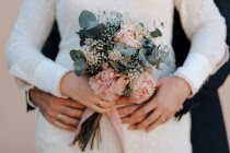 Crop anonymous groom embracing elegant bride in white wedding gown with delicate floral bouquet — Stock Photo