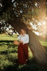 Jovem fêmea em blusa branca à moda antiga e saia segurando cesta de vime cheia de maçãs frescas e olhando para a câmera enquanto descansa perto da árvore no dia de verão no campo — Fotografia de Stock