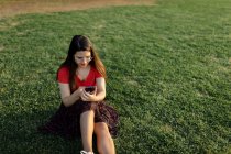 Weibchen in Sommerkleidung sitzen auf der grünen Wiese im Park und surfen mit dem Handy im Internet, während sie am Wochenende abends unterhalten werden — Stockfoto