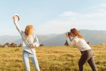 Side view female taking photo of blond girlfriend raising arm with hat spending time together in mountains — Stock Photo