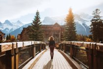 Rückansicht einer unkenntlichen Reisenden auf einer hölzernen Fußgängerbrücke vor einer bergigen Landschaft mit schneebedeckten Gipfeln, während sie Herbstferien in der Stadt Canmore in der Nähe des Banff-Nationalparks in Kanada verbringt — Stockfoto