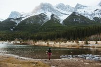 Vue arrière d'une randonneuse méconnaissable debout seule sur la rive du lac Quarry contre de majestueuses montagnes boisées avec des sommets rocheux enneigés dans le parc national Banff au Canada — Photo de stock
