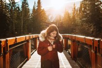 Jovem viajante feminina em outerwear desfrutando de um dia ensolarado de outono enquanto está de pé na ponte de madeira sobre o rio contra árvores e montanhas de coníferas no Parque Nacional Banff, no Canadá — Fotografia de Stock