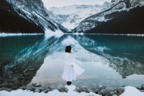Back view of anonymous female in white dress standing towards clean water of lake Louise against snowy mountain ridge on winter day in Alberta, Canada — Stock Photo