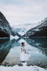 Vista lateral da fêmea em vestido branco e cachecol em pé em direção à água limpa do lago Louise contra cume de montanha nevado no dia de inverno em Alberta, Canadá — Fotografia de Stock