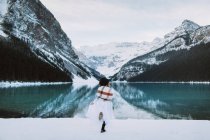 Vista posterior de la mujer anónima en vestido blanco y bufanda corriendo hacia el agua limpia del lago Louise contra la cresta de montaña nevada en el día de invierno en Alberta, Canadá - foto de stock