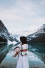 Rückansicht einer anonymen Frau in weißem Kleid und Schal, die an einem Wintertag in Alberta, Kanada, gegen den schneebedeckten Bergrücken am klaren Wasser des Lake Louise steht — Stockfoto